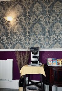 Innenaufnahme eines Hotels zeigt vor einer Ornament-Tapete eine Kaffeemaschine und Müslipäckchen.